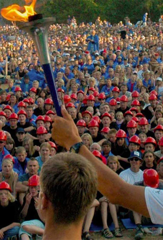 A stop on the Sri Chinmoy Peace run is pictured with the burning peace torch held up in the hands of an organiser with his back turned. Opposite him, a huge crowd of thousands of people have gathered to be part of the Sri Chinmoy Peace Run.