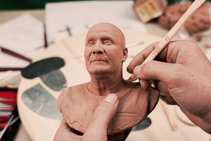 The sculptor holds the bust in his hand and shapes it out of clay using a wooden tool. Sculpting tools and sculpting clay are slightly blurred in the background with some sketches on a table. The clay sculpture in the making shows the early artistic phase of the bronze bust as the sketches take shape.