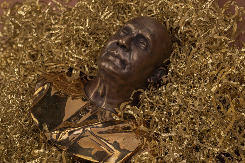 A shimmering gold polished bronze bust lying on a bed of thick golden copper chips, expressing the fact that the bronze material used is completely recycled.