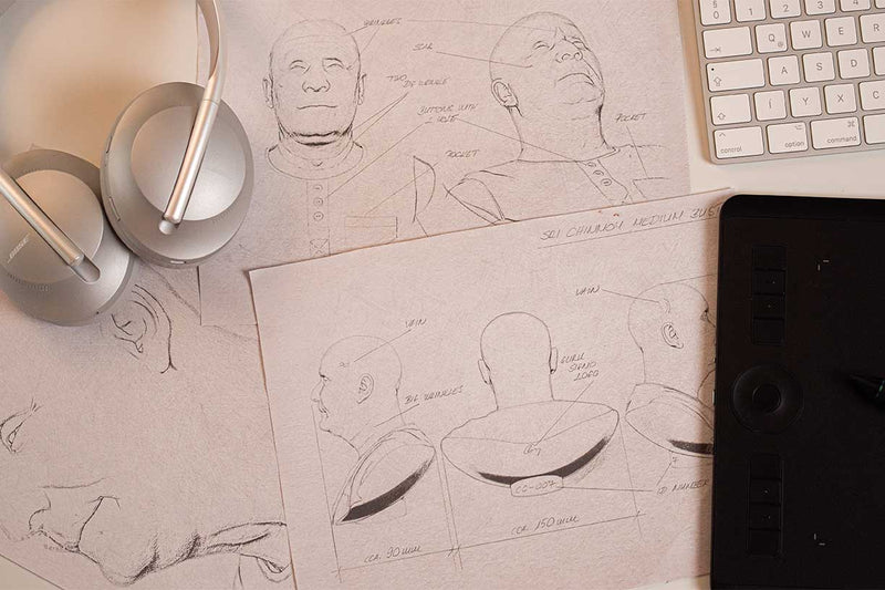 The picture shows the plans of the bronze sculpture of Sri Chinmoy drawn on paper in various angles. The pencil drawing has markings and handwritten annotations. In addition to the pencil drawings on paper, there is also a Bose headset and a part of an Apple Magic Keyboard.