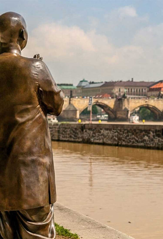 One of the bronze statues of Sri Chinmoy in Prague. The statue stands on the river bank, with the city centre in the background.