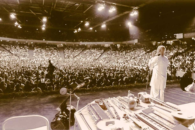Sri Chinmoy gives a free peace concert for thousands of people. He is pictured standing on stage praying in front of the audience.